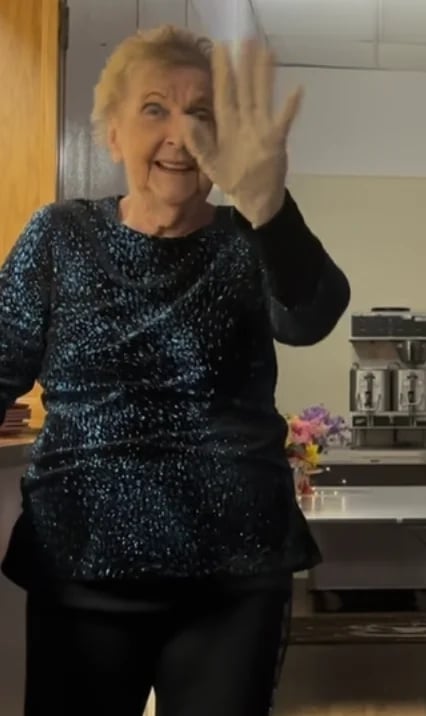 Older woman with blonde hair wearing a sparkly dark top waving and smiling in an indoor setting