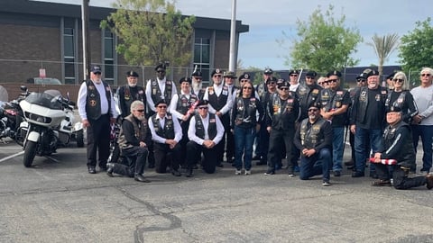 Large group of people in leather vests and black clothing posing for a photo with a motorcycle, outdoors on a parking lot