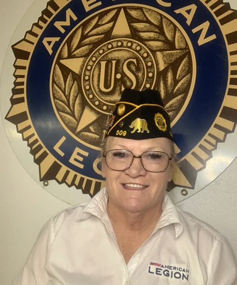 Smiling woman wearing glasses and American Legion cap in front of the American Legion seal emblem