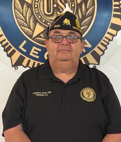 Man in black polo shirt with military insignia wearing glasses and cap, standing in front of official seal backdrop