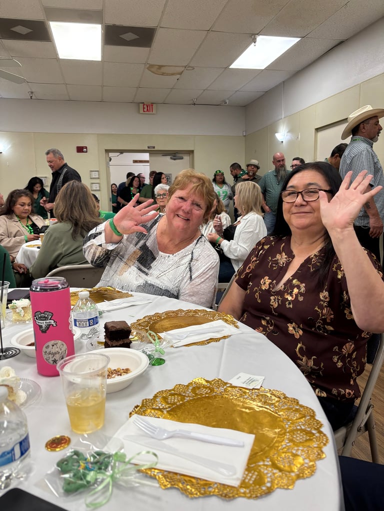 Two women waving at a crowded community dinner event in a cafeteria with tables of food and many guests in the background