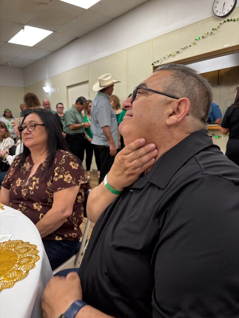 Man in black shirt and glasses listening thoughtfully at an indoor community event with people gathered in background