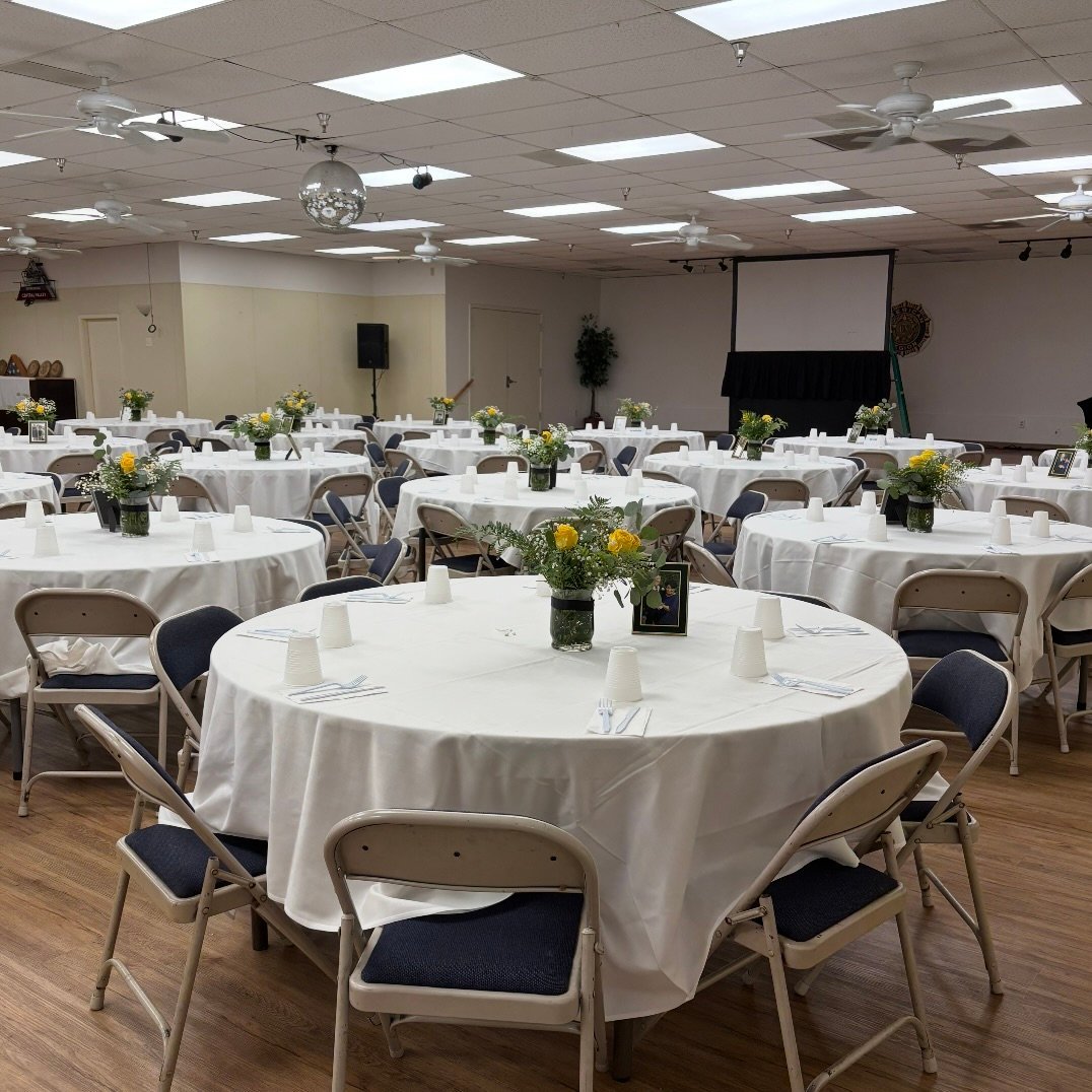 Event venue with round tables covered in white tablecloths, yellow flowers, and place settings arranged for a formal dinner or celebration
