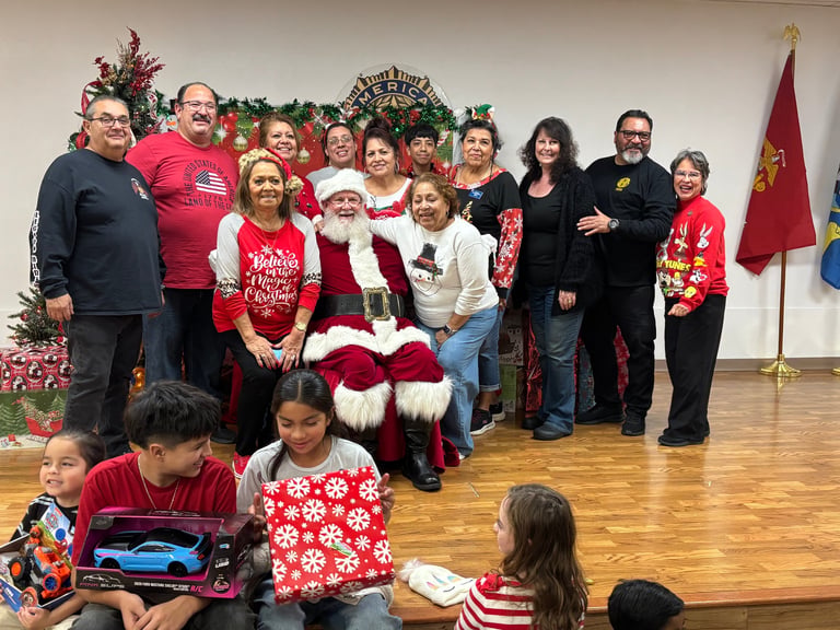 Large group posing with Santa Claus indoors, including adults and children in holiday attire, with a red flag visible in background