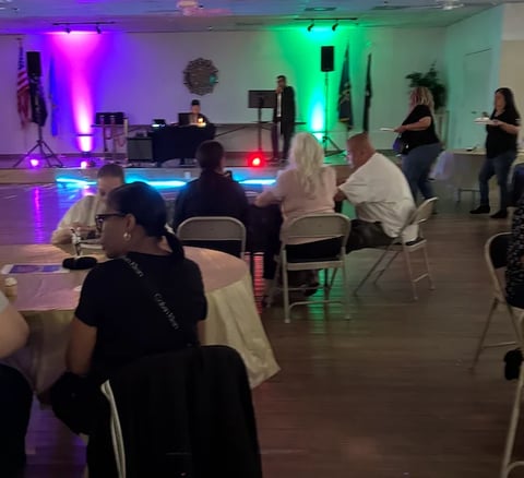 Indoor event space with attendees seated and standing, DJ booth with colorful purple and green neon lighting, dance floor, and audio equipment