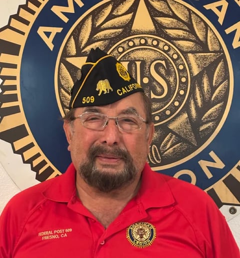 Man wearing red American Legion shirt and military cap with California insignia, standing in front of gold and blue emblem