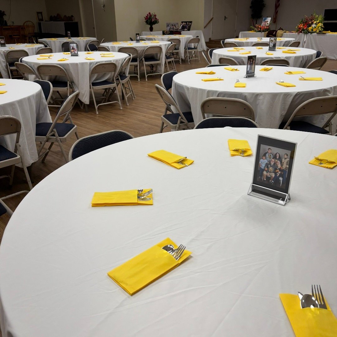 Banquet hall with round tables covered in white tablecloths, yellow napkins, place settings, and small framed photos as centerpieces