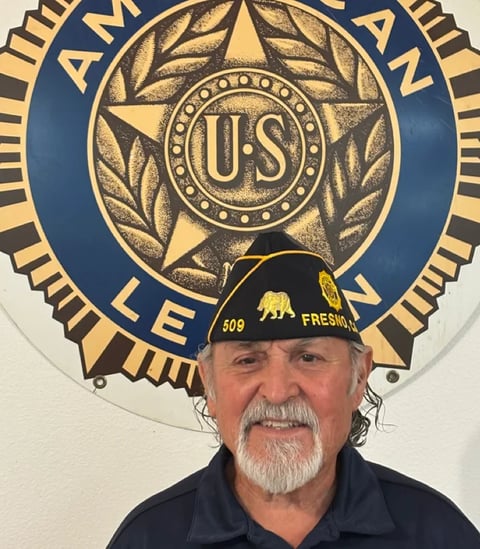 Man with gray beard wearing a black cap and dark shirt, smiling in front of American Legion emblem with gold star and wreath design