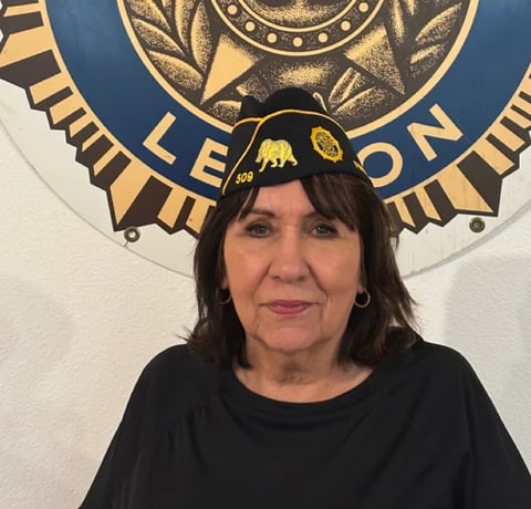 Woman with dark hair and bangs wearing a black shirt, positioned in front of a decorative blue and gold seal emblem on a white wall