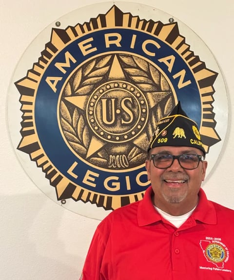 Man in red American Legion polo shirt and military cap smiling in front of American Legion emblem on wall