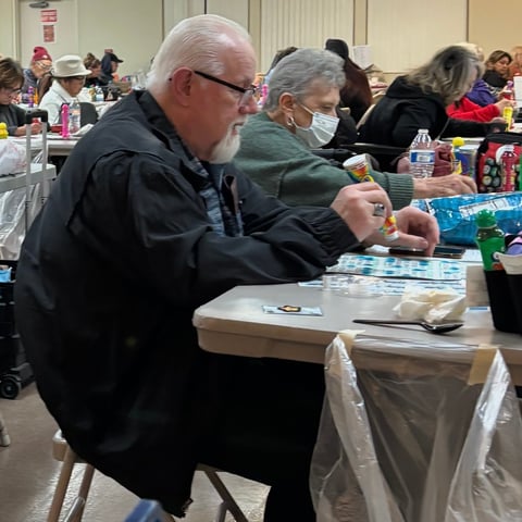 Two men seated at a table at an indoor gathering, examining items displayed in front of them, with other attendees visible in the background