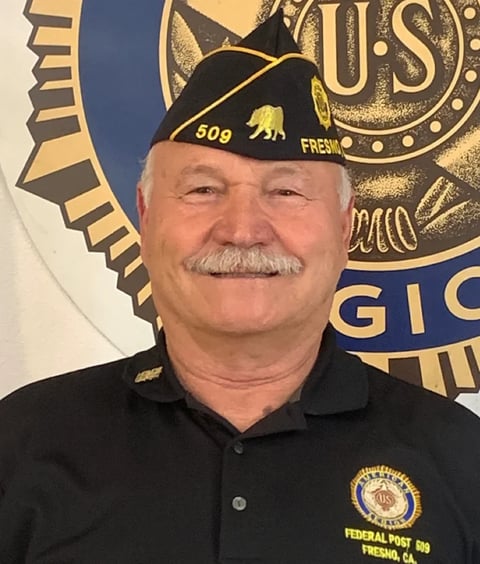 Older man with gray mustache wearing black VFW hat and shirt with emblem, standing in front of American Legion banner