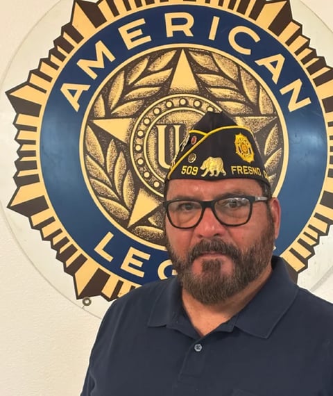 Man with beard and glasses wearing a black cap in front of American Legion emblem on wall