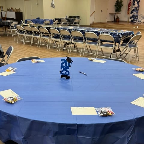 A blue round table with place settings in an event space with rows of blue and tan folding chairs arranged in the background