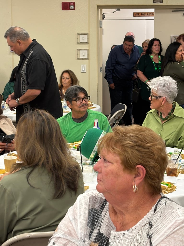 Community gathering in an indoor room with diverse adults seated and standing around tables during what appears to be a social event or meal
