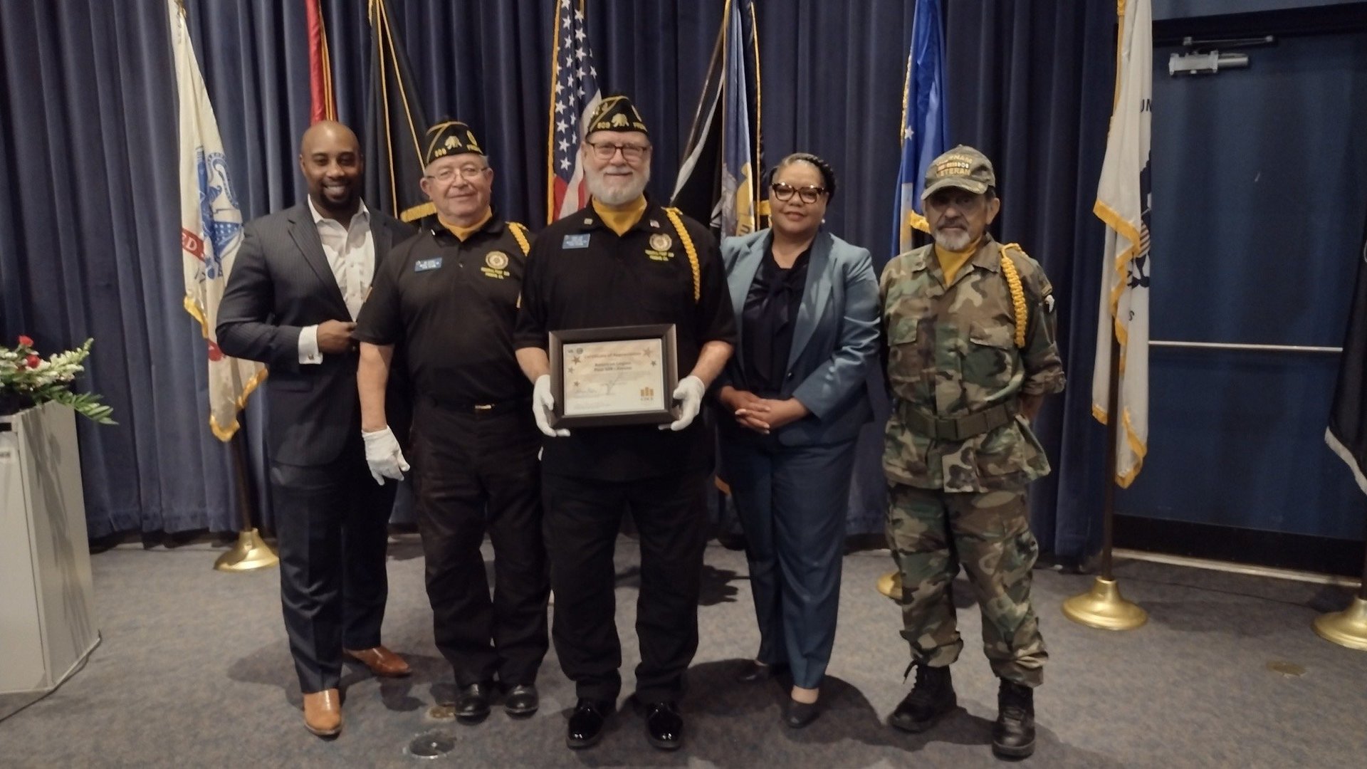 Five people standing in formal attire and military uniforms holding a certificate in front of flags and blue curtains
