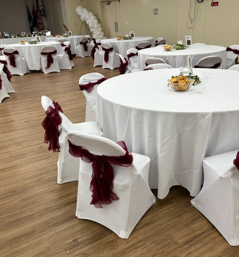 Elegant event venue setup with white round tables covered in white tablecloths, decorated with burgundy fabric sashes on white chair covers, and white balloon arch in background