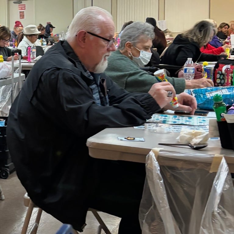 An older man with glasses sits at a table with others at what appears to be a crowded indoor community event or gathering