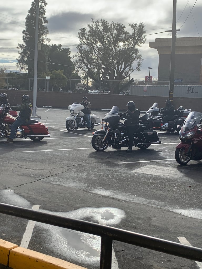 Group of motorcyclists and riders gathered in a parking lot on an overcast day with industrial buildings in the background