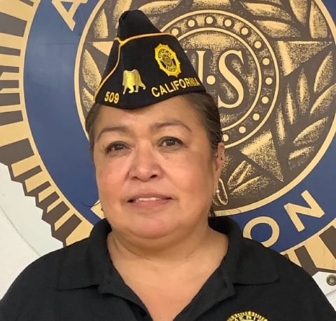 Woman wearing black polo shirt and California VFW hat, posing in front of blue and gold VFW emblem backdrop