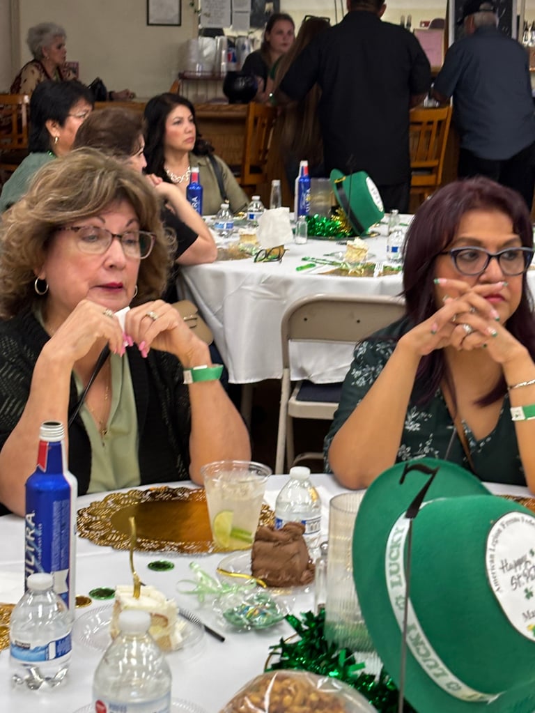 Two women at a formal event seated at a table with green decorations and place settings, with other attendees visible in the background