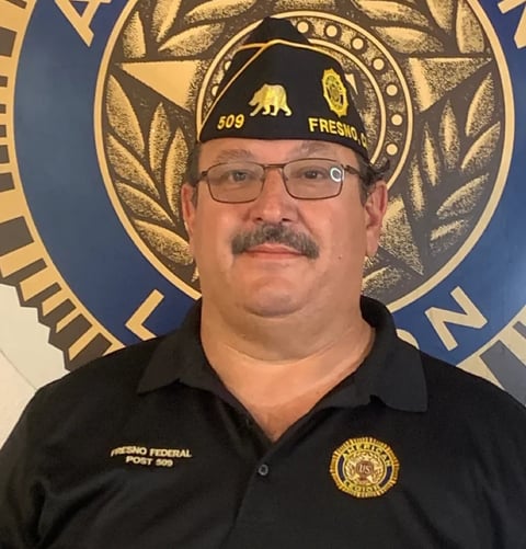 Man with mustache wearing glasses and black Fresno Federal Post 509 shirt in front of emblem