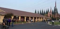 Outdoor marketplace or courtyard with colonial-style buildings, visitors shopping, and evergreen trees in background