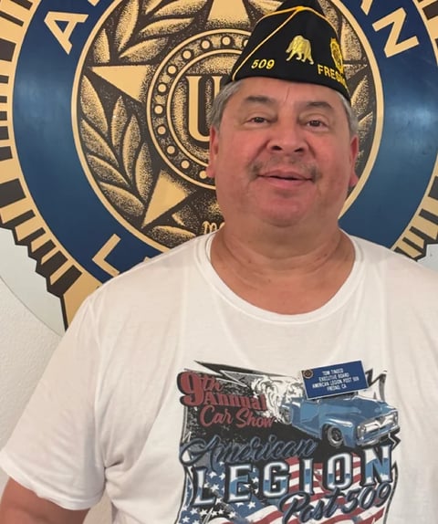 Middle-aged man wearing a white t-shirt and military-style cap with #409 insignia, smiling at camera in front of an American Legion emblem