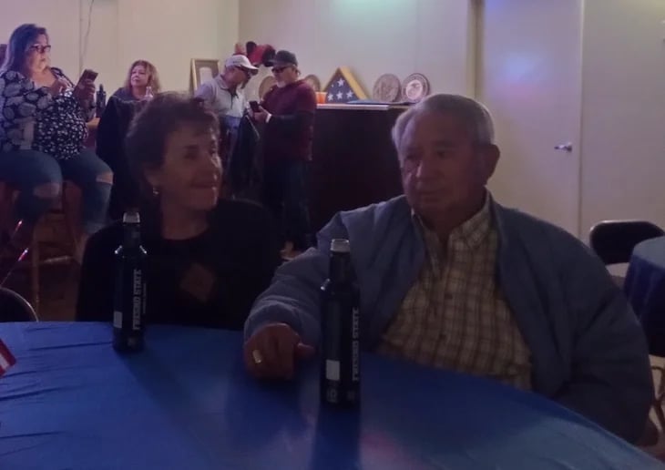 Two people seated at a blue table facing each other during an indoor event with other attendees visible in the background