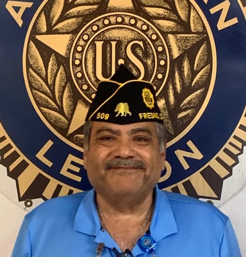 Man with mustache in blue shirt smiling in front of official seal or emblem with gold and blue colors