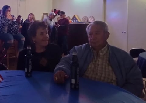 Two people seated at a blue table during an indoor event, with other attendees visible in the background