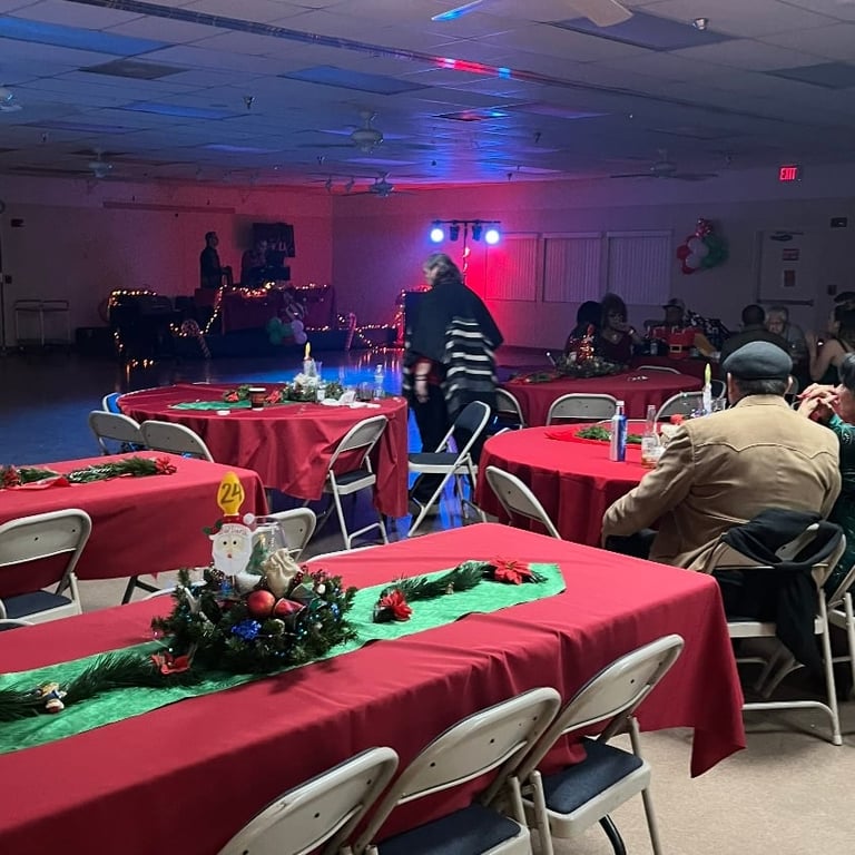 Indoor event space with red-draped tables, colorful blue and pink lighting, DJ booth, and guests seated at tables during a celebration