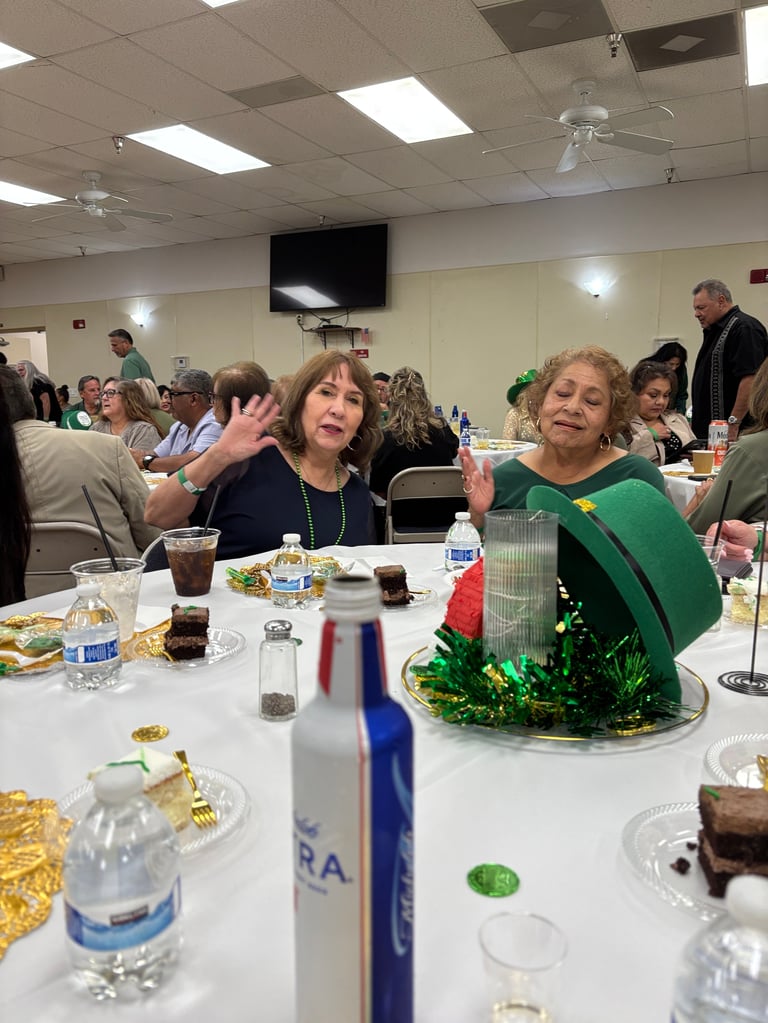 Two women waving at a dining table during a community event in an indoor venue with food and decorations