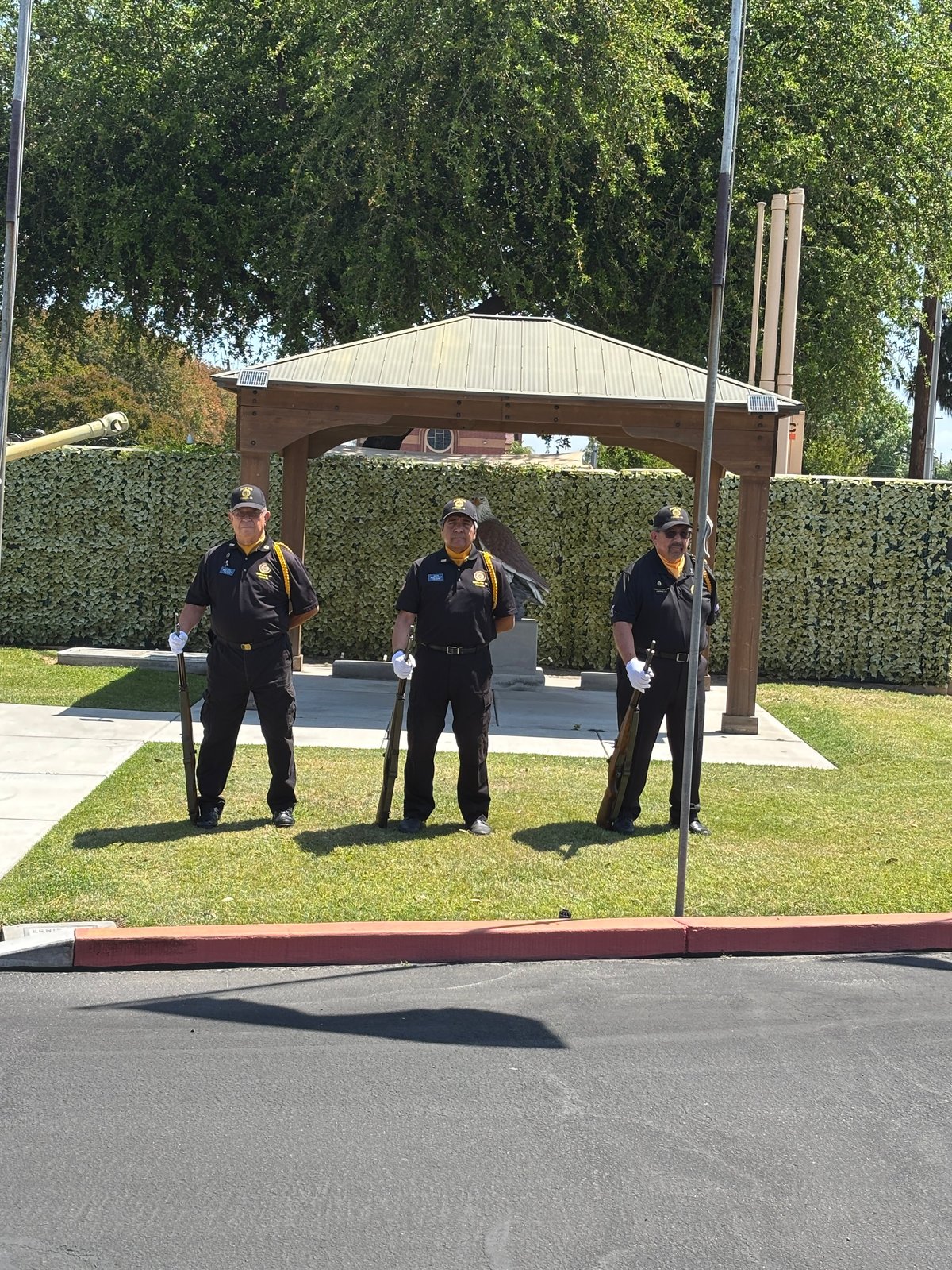 Three uniformed officers stand on grass beneath a wooden shelter structure with a metal roof at a public facility.