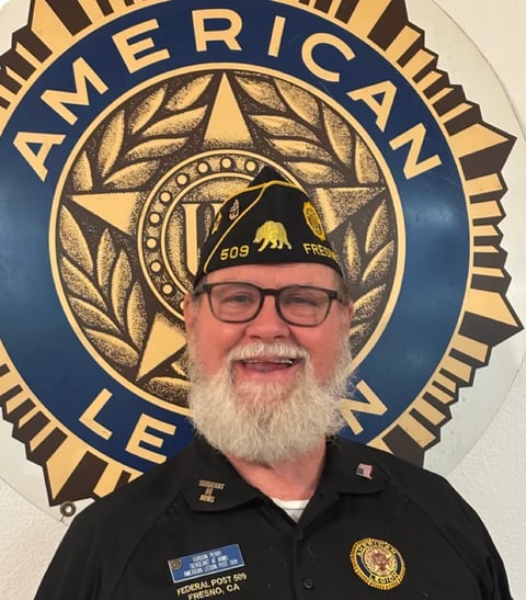 American Legion member wearing glasses and cap smiling in front of blue and gold legion emblem