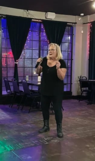 Woman with blonde hair singing into a microphone on a stage with purple lighting and large windows behind her